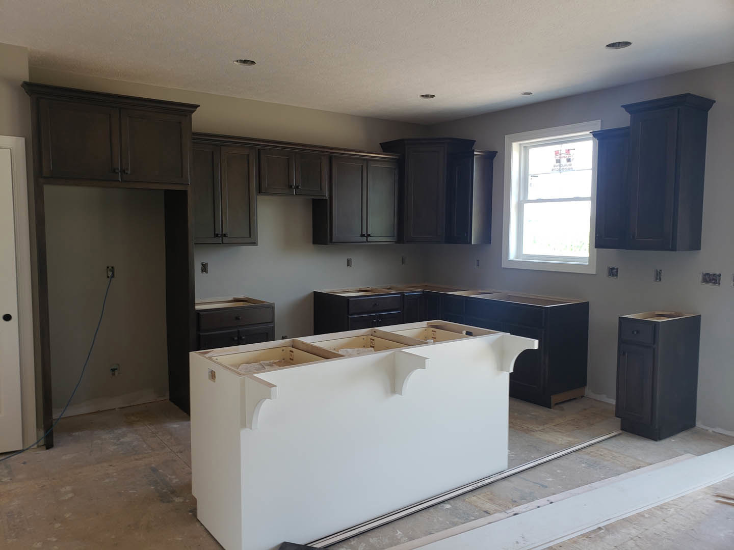 Kitchen featuring dark wood cabinets, white island with quartz countertop and open shelving, stainless steel sink, black drawer unit, window with sign, blue electrical cord on
