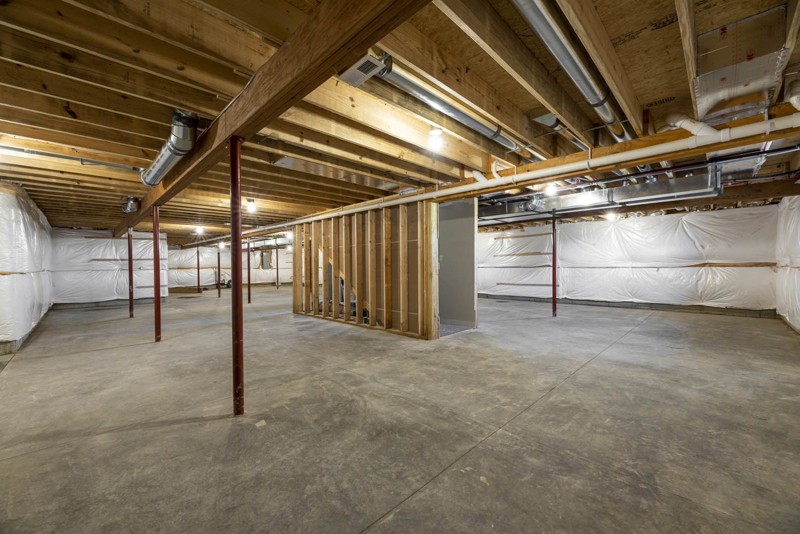 Exposed wood ceiling beams, concrete floor, white plastic sheet covering part of a wall, white door with wooden frame, steel pole in unfinished basement room