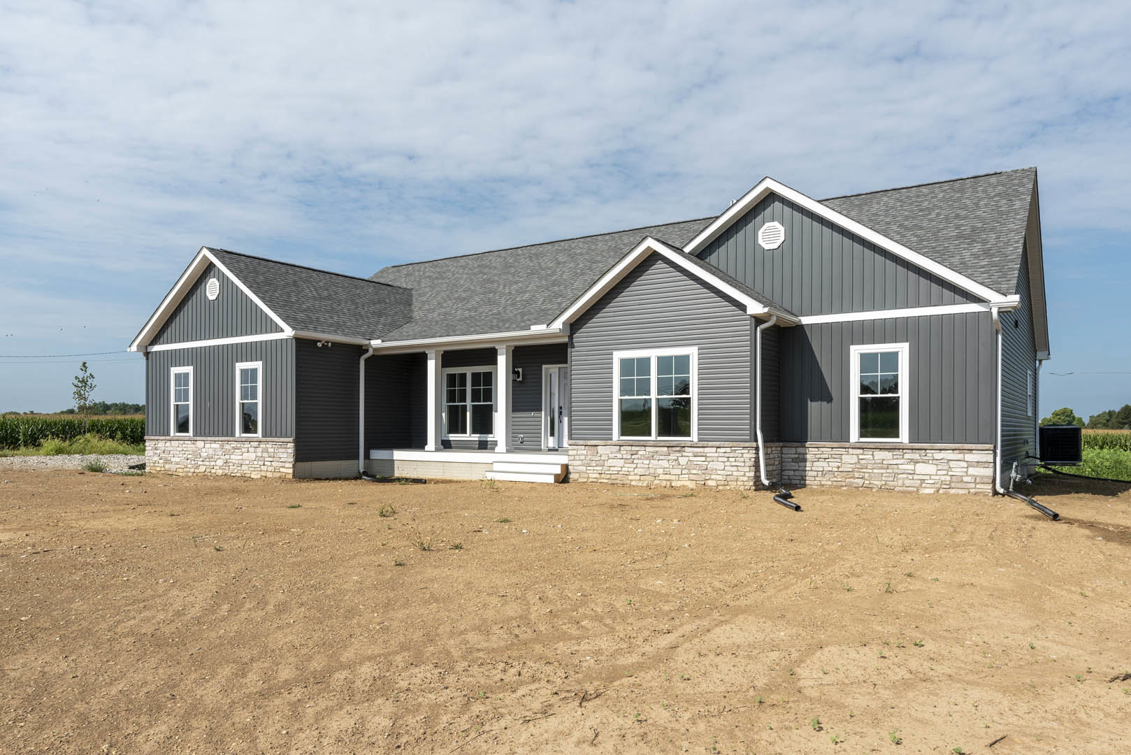 White house with a metal roof and white-framed windows, stone retaining wall bordering a dirt field, under a cloudy sky