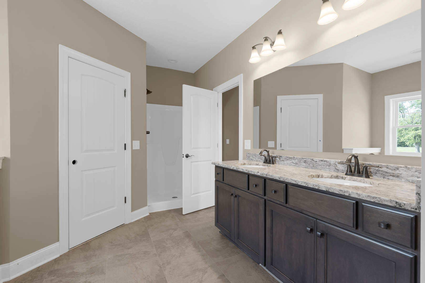 Bathroom with double vanity featuring white countertops and black faucets, glass-enclosed shower with tile walls, white door with black handle, window overlooking trees