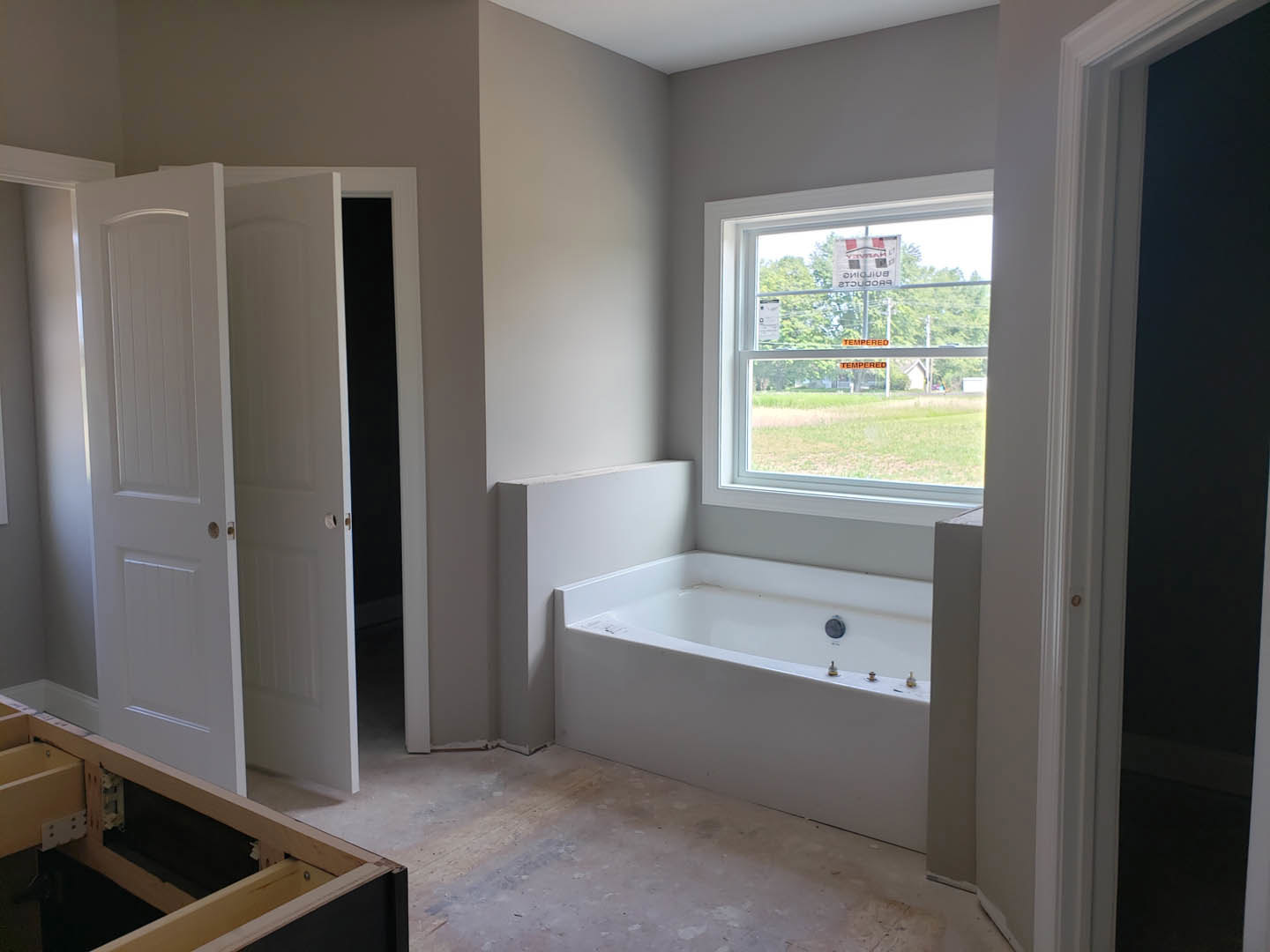 White freestanding bathtub beside a large window, wooden shelf mounted on wall, white door with black handle, neutral cabinetry and light-colored walls