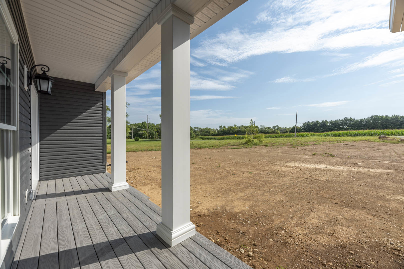 Covered porch with white pillars, wooden deck, and adjacent dirt field bordered by grass and trees under a partly cloudy blue sky