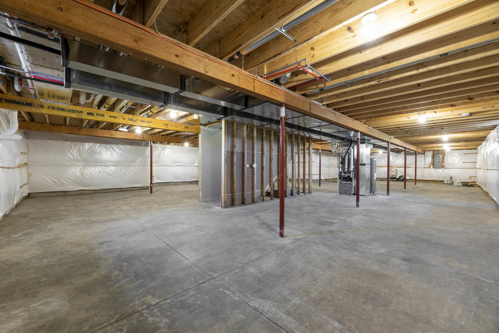 Exposed wood and metal beams across ceiling, concrete floor with red support pole, visible pipes and grey metal box with white label, white plastic covering on wall in unfinished