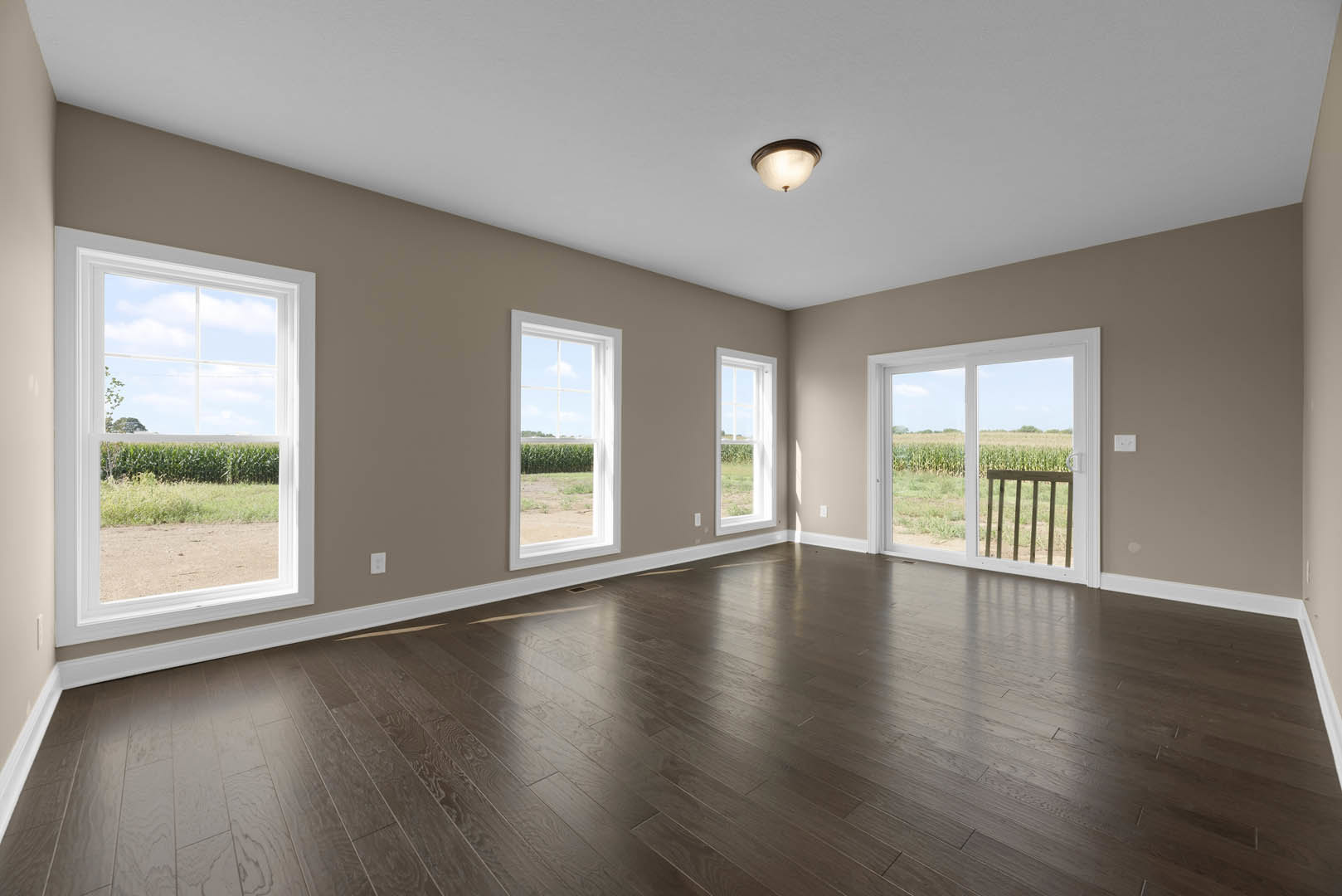 Dark wood floor in a spacious room with large windows and sliding glass door overlooking a corn field, ceiling light fixture, and wooden railing.