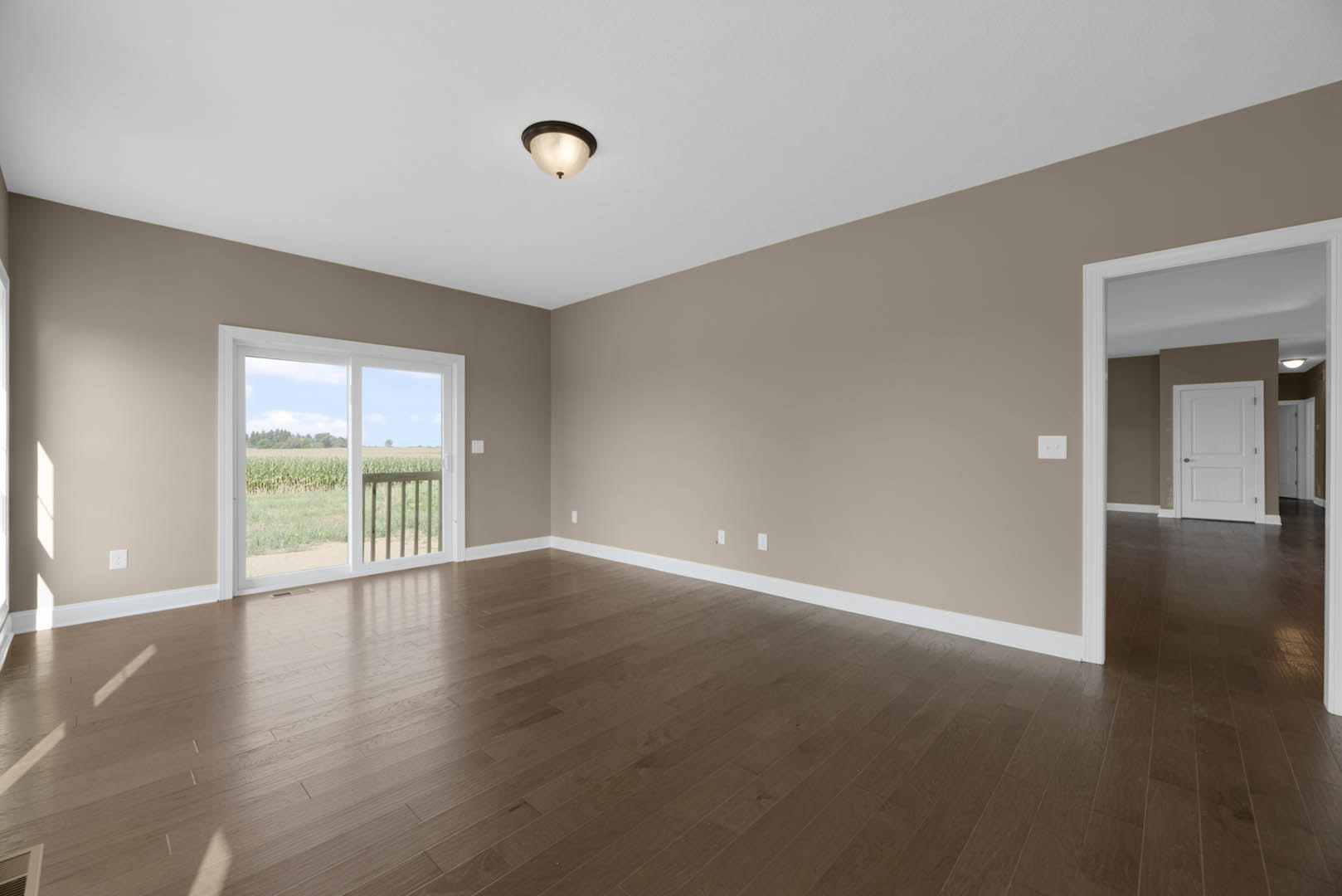 Room with wood laminate flooring, white plaster walls, white door with silver knobs, sliding glass door opening to balcony and view of cornfield, ceiling light fixture