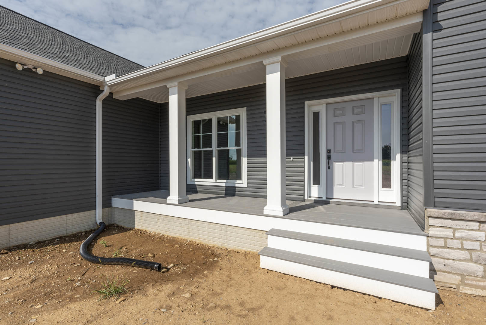White front door with glass panes, black pipe lying on dirt near porch, gray siding, exterior window, and partial view of roof and staircase step