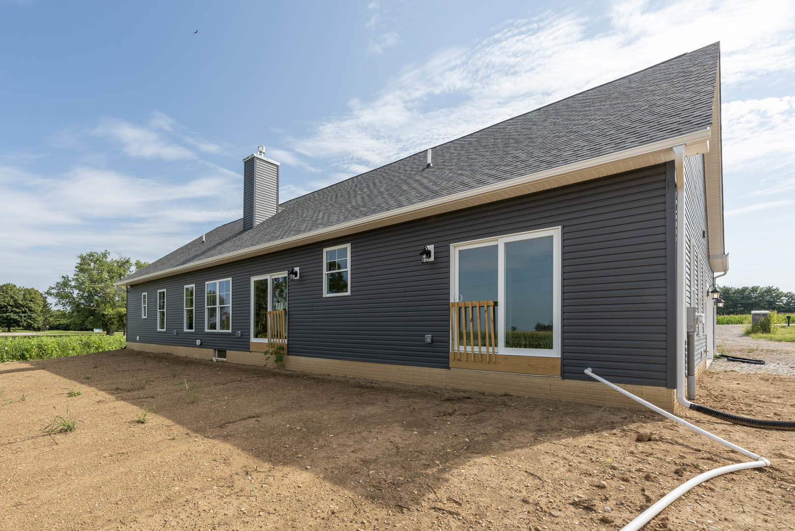 Two-story home with beige siding, white-framed windows, and a white railing, set on a dirt yard bordered by a wooden fence, under a partly cloudy sky.