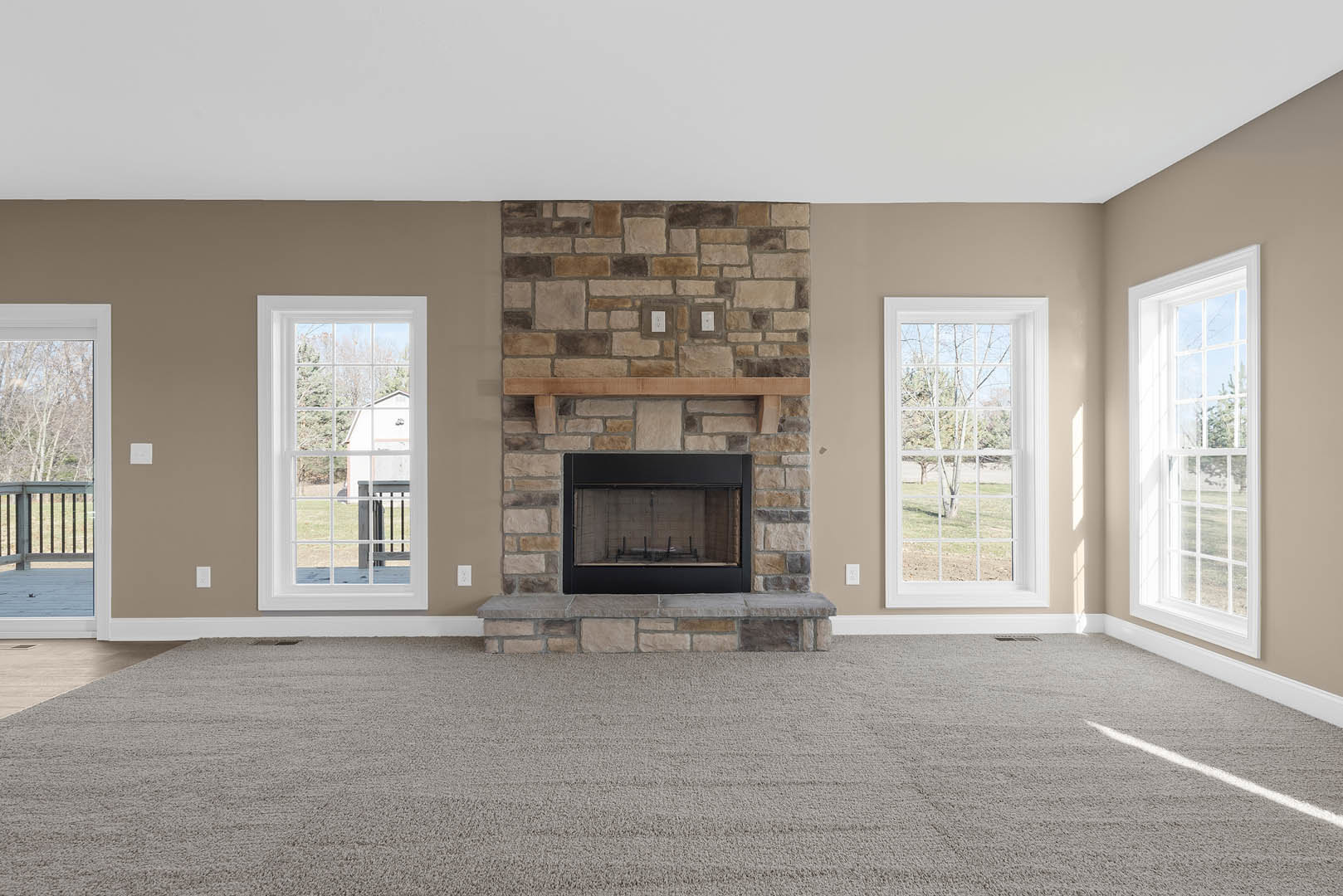 Stone fireplace with glass door set in a den, hardwood flooring, large windows showing trees and neighboring house under cloudy sky