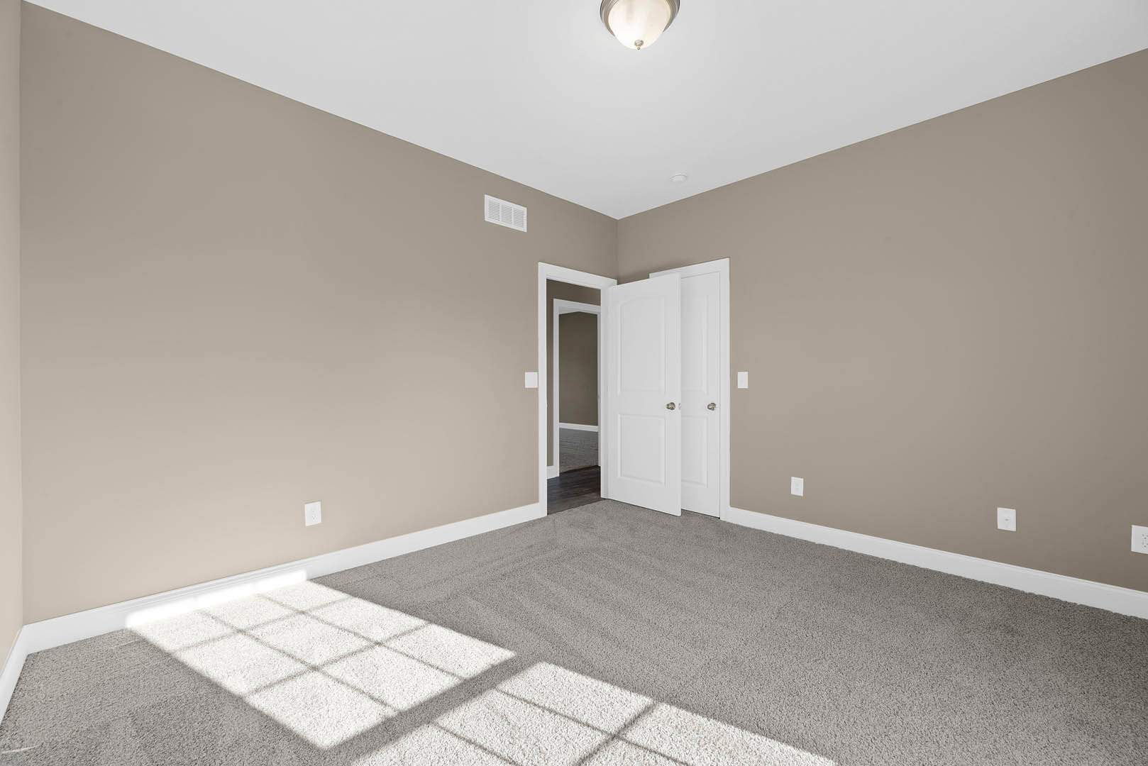 White paneled door with silver knobs, beige carpeted floor featuring a white rug, recessed ceiling light, wall vent, and white baseboard molding in a neutral-toned room.