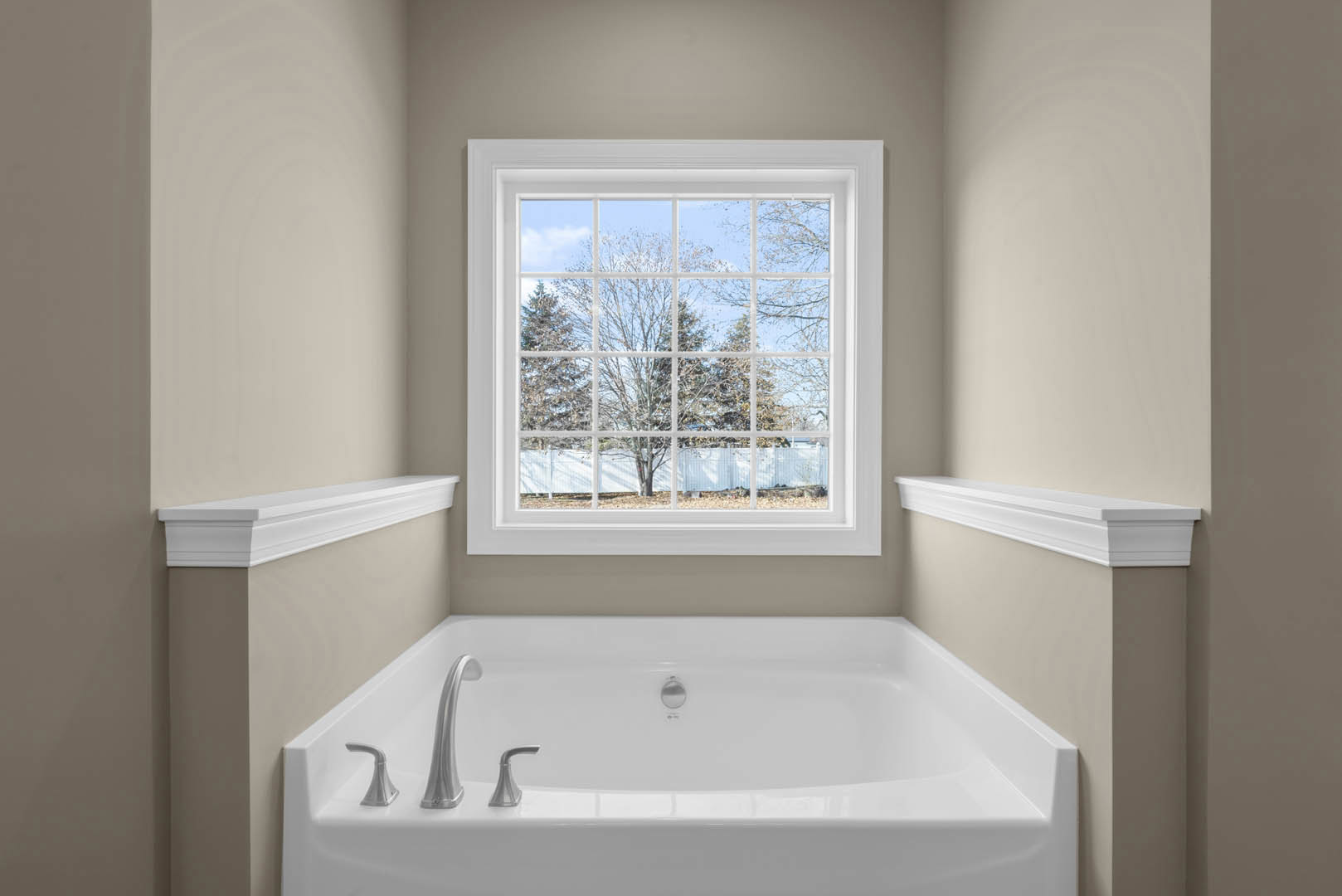 Freestanding white bathtub beneath large window with view of trees and fence, white wall molding and shelf, chrome faucet, bright bathroom interior