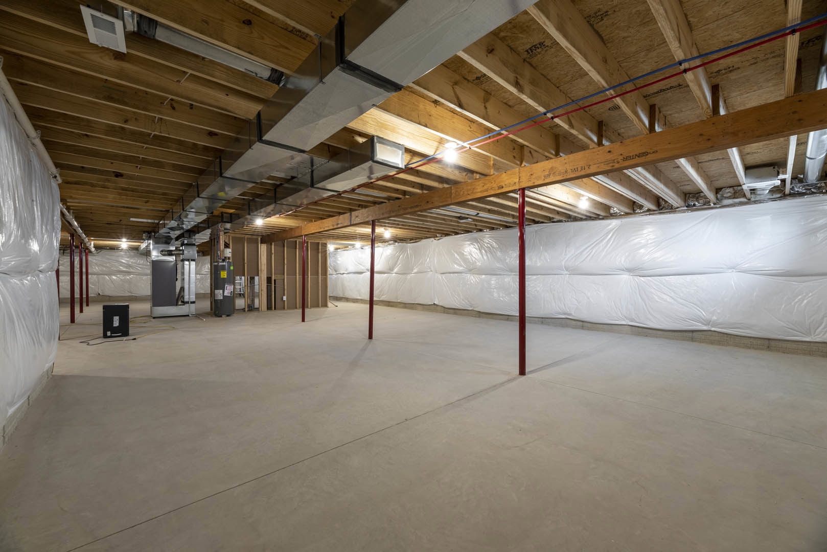 Exposed wooden ceiling beams, white wall partially covered with plastic sheeting, concrete floor with red steel poles, and scattered white plastic bags in an unfinished basement