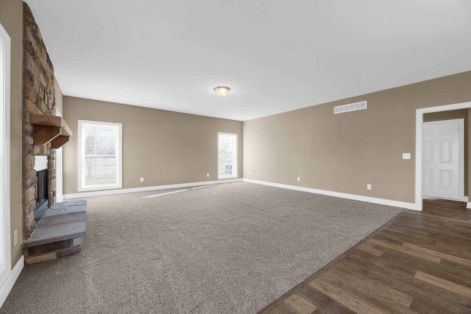Carpeted living room with a stone fireplace, white door with brown trim, large window overlooking trees, and ceiling light fixture