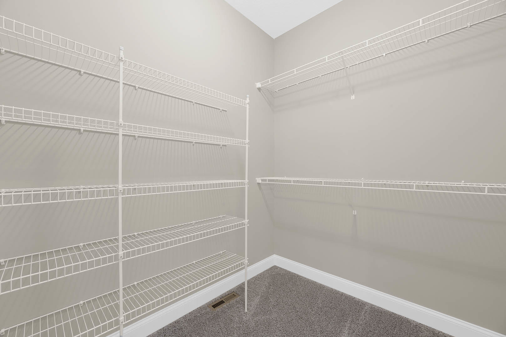 White wire shelves mounted on a wall above beige carpet with a floor vent, white ceiling, and smooth plaster walls.