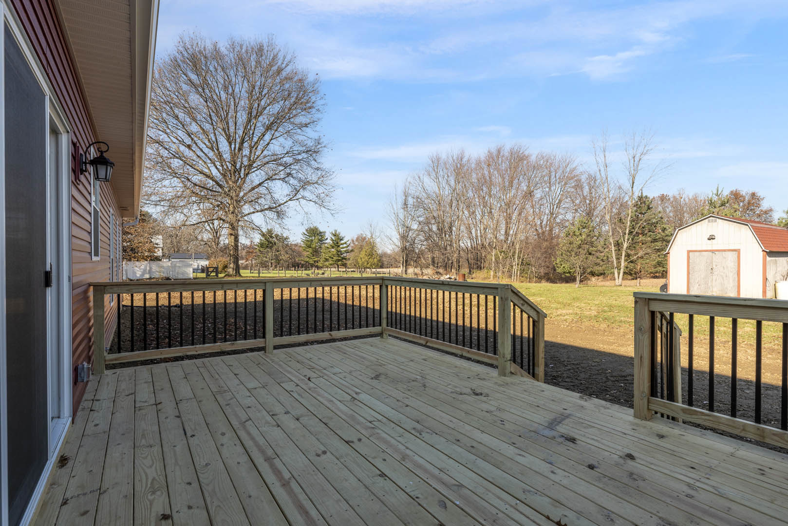 Wooden deck with black metal railing, fenced yard, leafless tree, and white shed with red roof in the background.