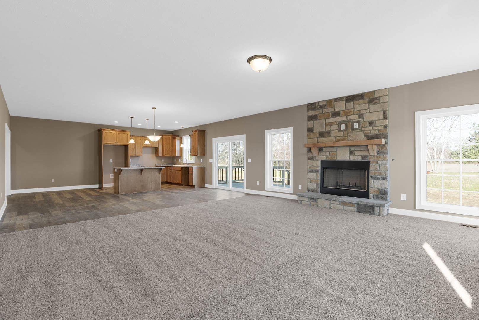Open living room with wood flooring, carpeted area, glass-door fireplace, large multi-pane window with exterior railing, recessed ceiling lights, and adjacent kitchen space.