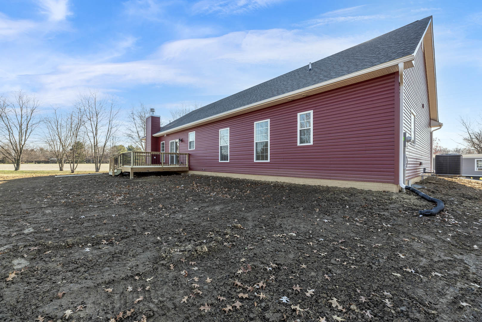 Red horizontal siding exterior with white-trimmed multi-pane windows, wooden deck with railing, dirt yard scattered with leaves, mature trees in background.