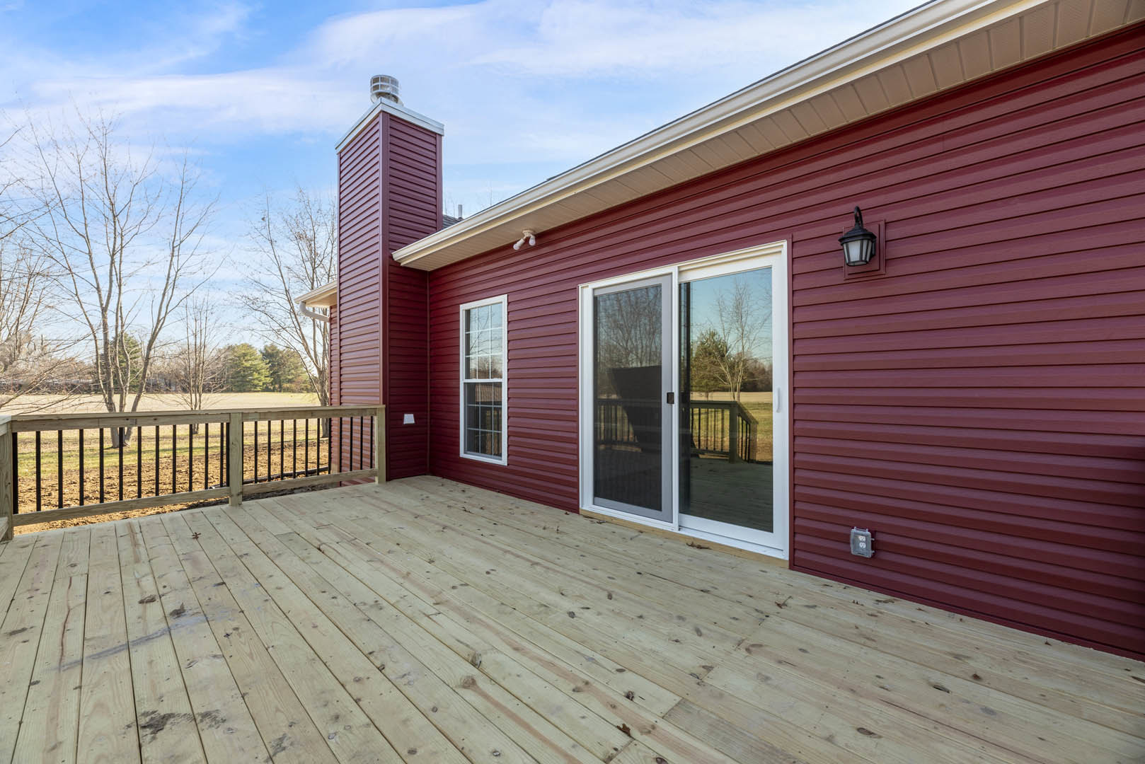 Red siding exterior with brick chimney, wooden deck featuring black railings, sliding glass door overlooking green lawn and trees, adjacent window, close-up of outdoor lamp and