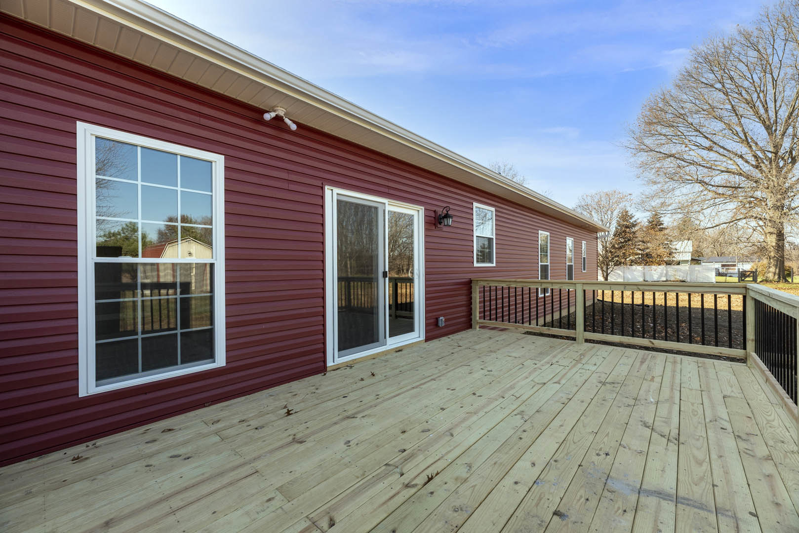 Wooden deck with railing and fence, red siding house with white-framed window and sliding glass door, leafless tree in the yard, cloudy sky overhead