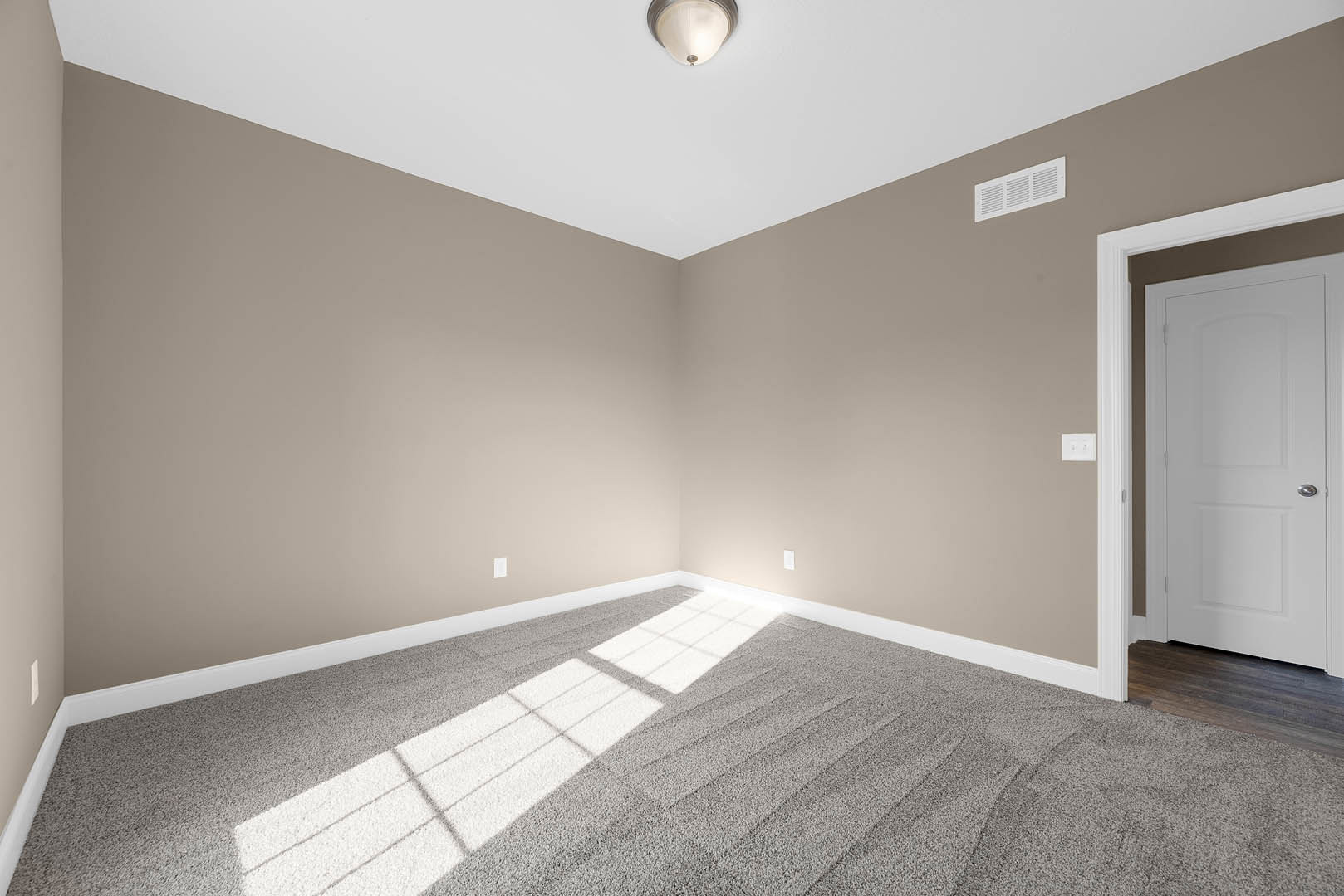 Carpeted room with white walls, ceiling light fixture, white door with silver handle, ceiling vent, and sunlight streaming through window onto floor