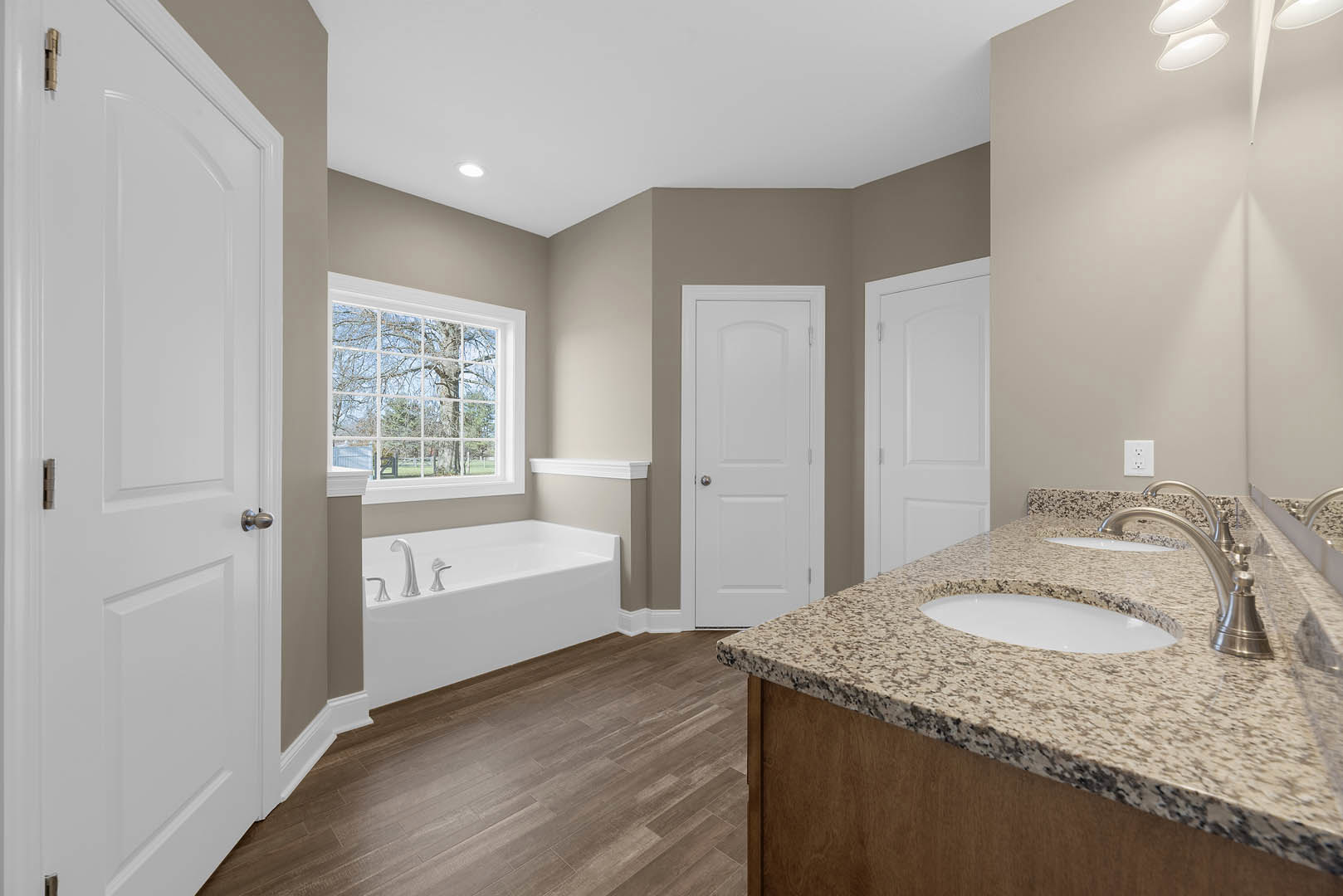 Bathroom with marble countertop and dual sinks, freestanding marble tub, wood flooring, white door with silver knob, large window with multiple panes