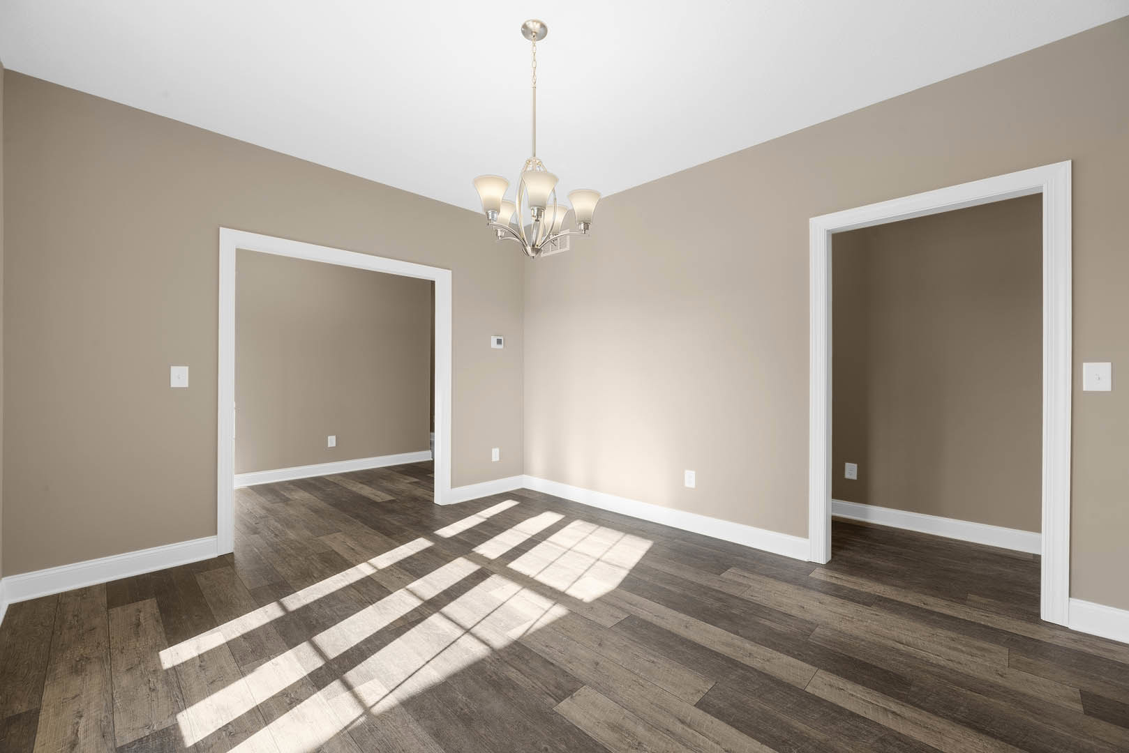 Chandelier hanging from white ceiling above wood flooring, natural light streaming through window, white walls with decorative molding, doorway with white trim visible