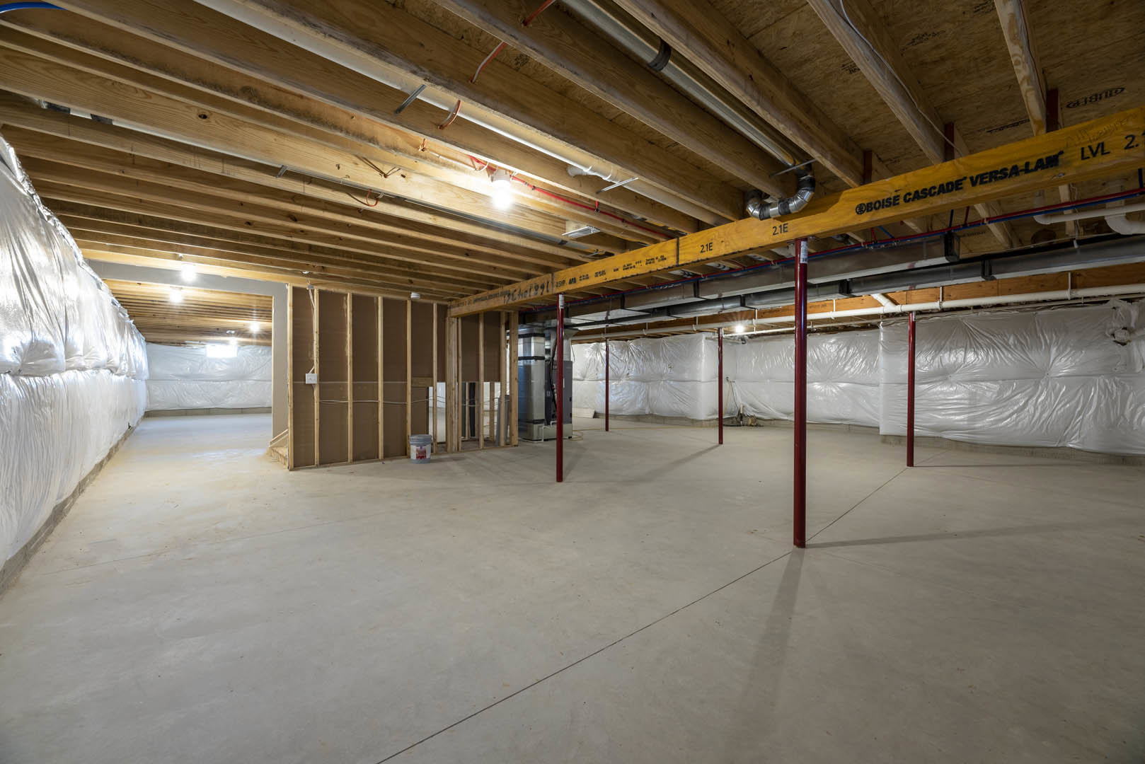 Basement room with exposed ceiling beams and pipes, concrete floor with red steel support pole, white plastic sheeting covering one wall, overhead lights, and visible composite