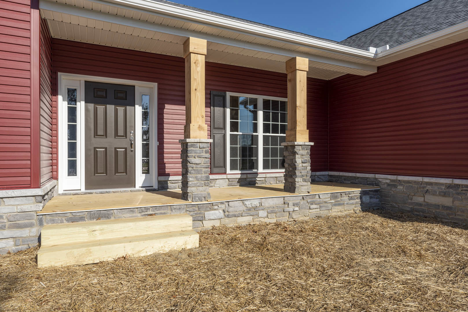 Red exterior wall with grey door, brown door featuring glass panes, white-framed window, wood siding, stone pillar, and pile of hay on porch.