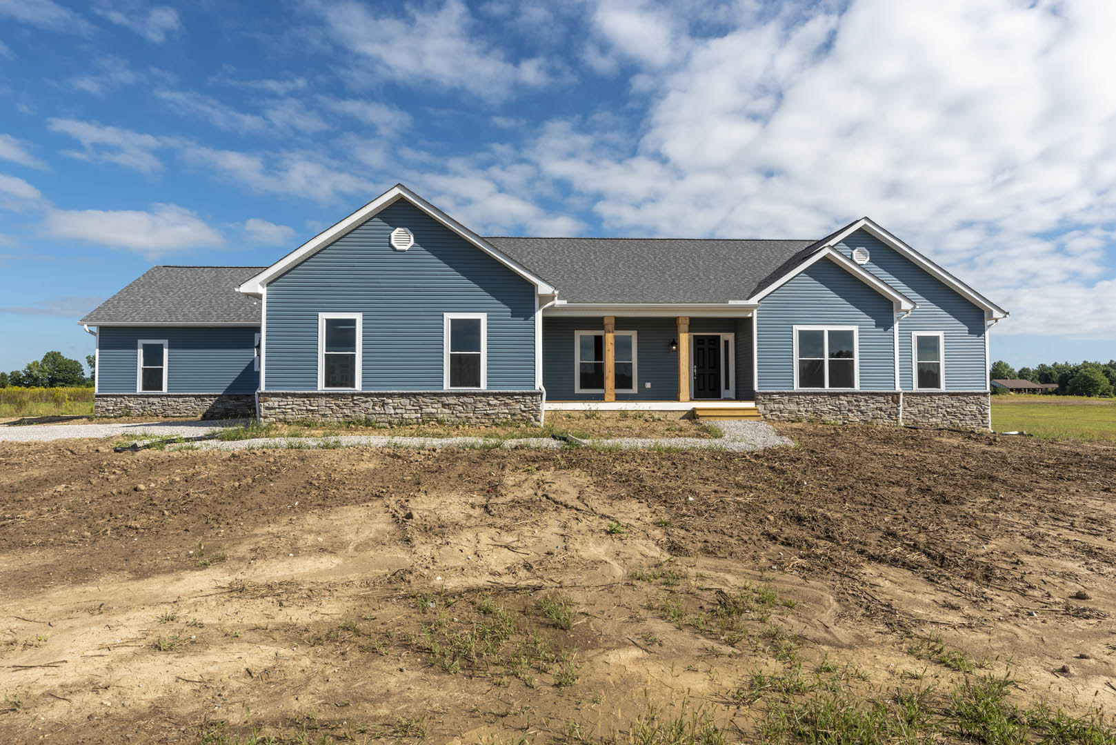 Two-story house with blue roof, white-framed window, black door with white trim, stone wall, surrounded by dirt hill and patchy grass under cloudy sky