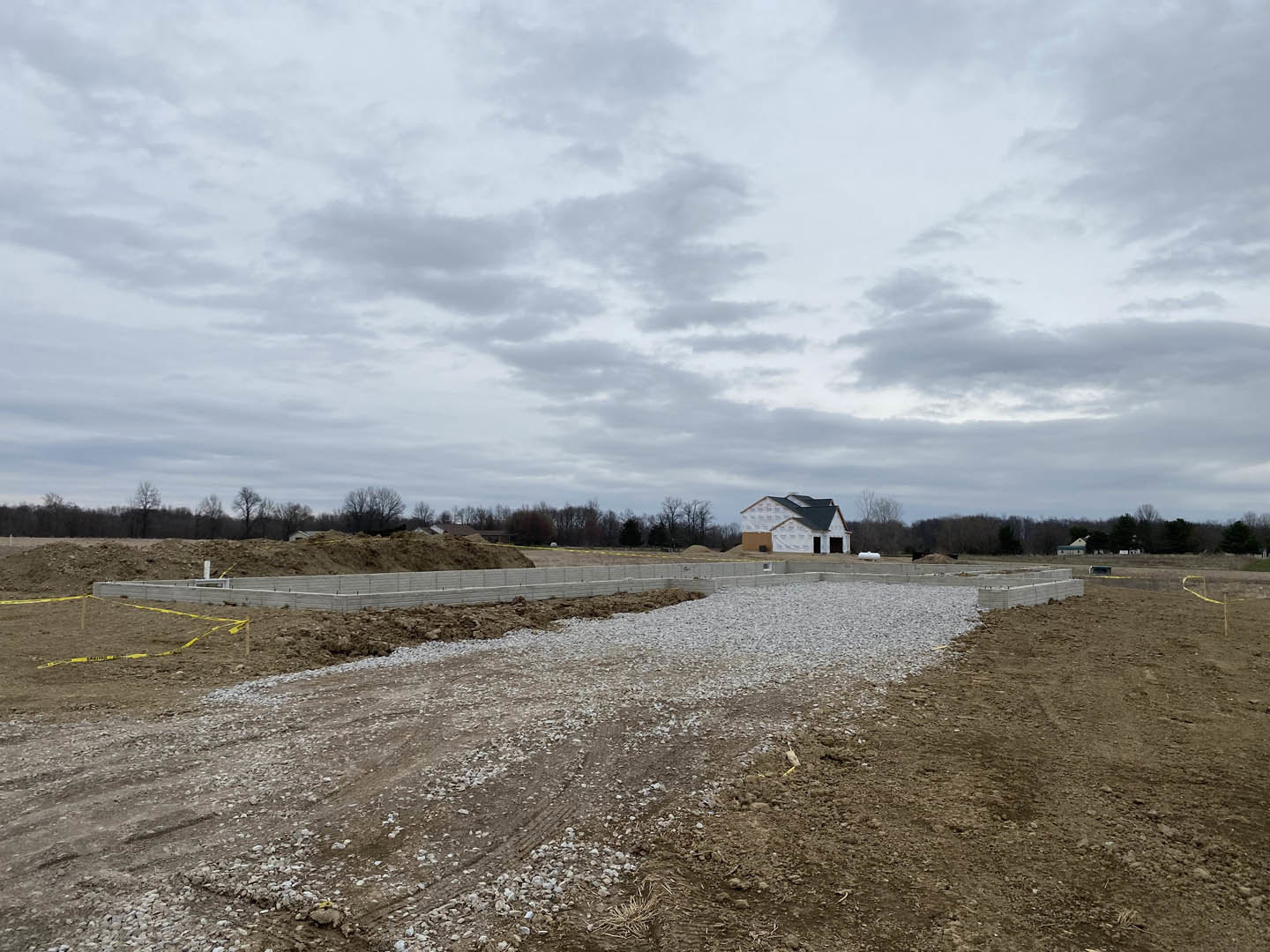 Gravel dirt road bordered by a low wall and yellow tape, leading to a white house with a black roof set against a cloudy sky and grassy field.