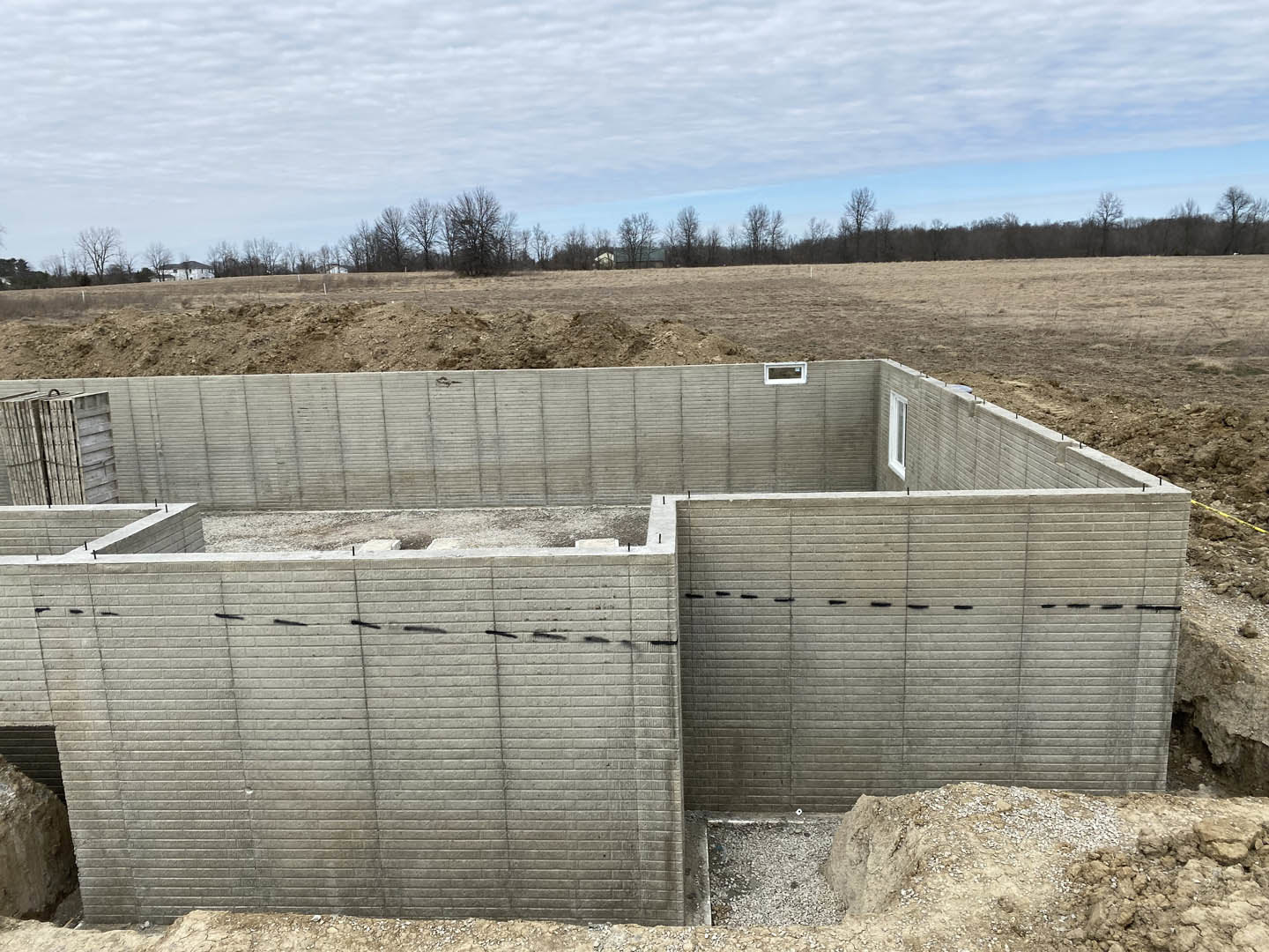 Concrete foundation wall with black tape accents set in a grassy field, cloudy sky and trees in background, stack of wooden crates nearby.
