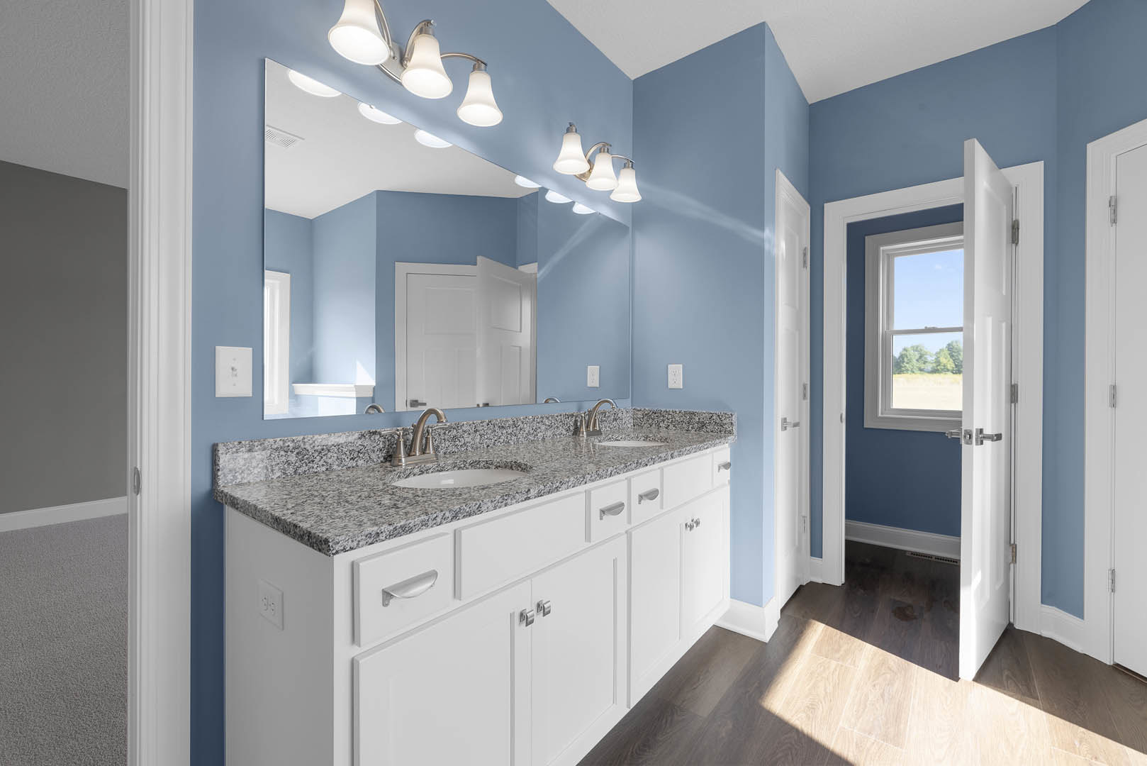 Bathroom with granite countertop sink, large wall mirror, white walls, silver door handle, ceiling light fixture, and window overlooking trees.