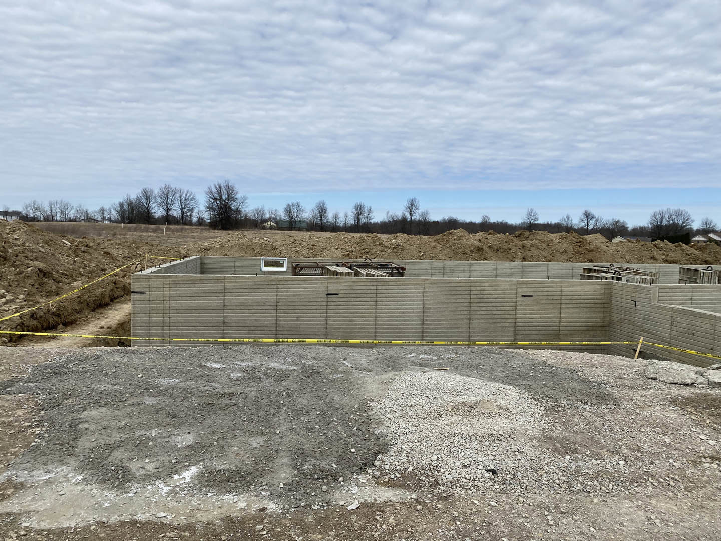 Expansive dirt construction site bordered by a concrete wall marked with yellow tape, scattered gravel piles, leafless tree, and surrounding wooded area under a cloudy sky