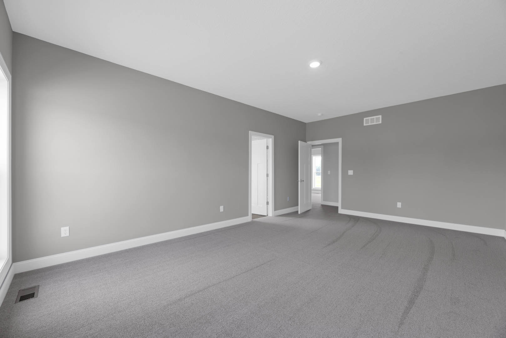 Grey carpeted room with light grey walls, open white door, white window trim, wall vent, and ceiling.
