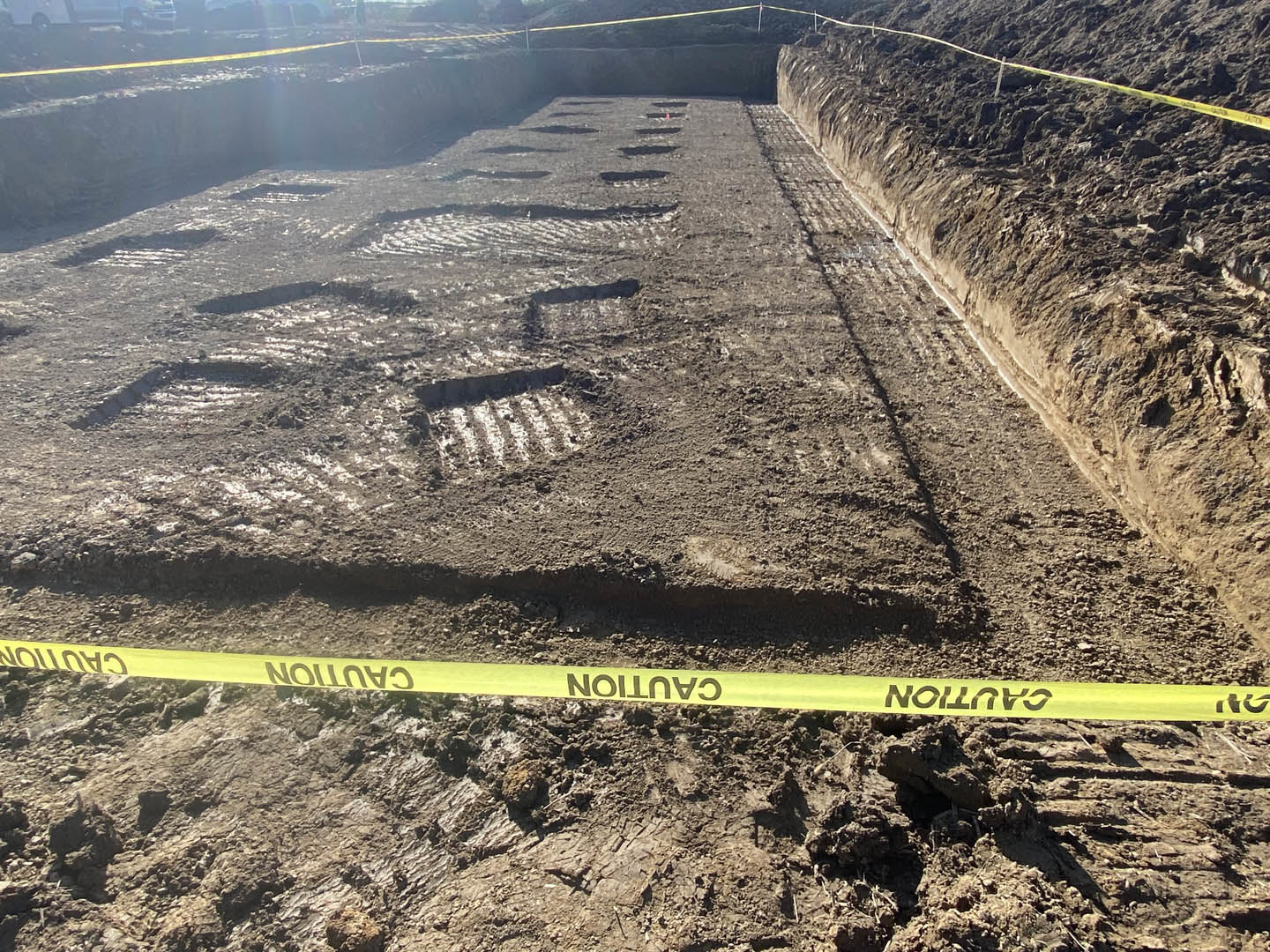 Yellow caution tape stretched across dirt at a residential construction site, with a yellow sign displaying black numbers and letters, and a man standing in the background.