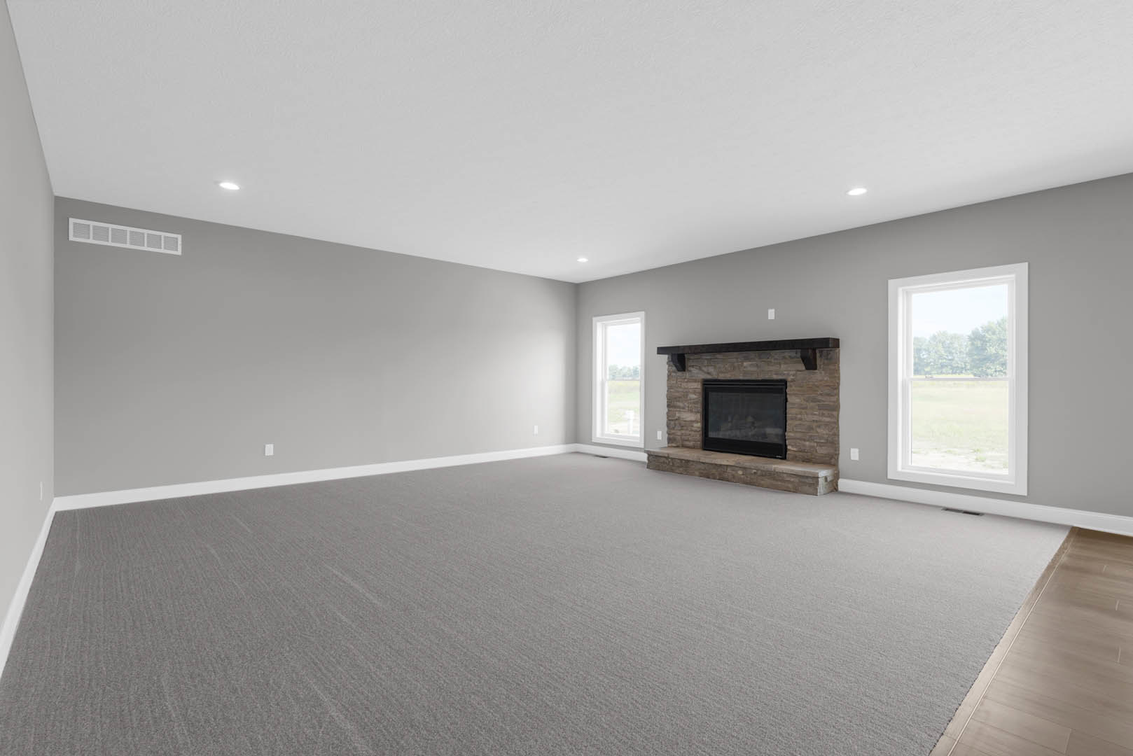 Living room with black-framed fireplace, large windows overlooking trees and field, carpeted floor, and staircase.