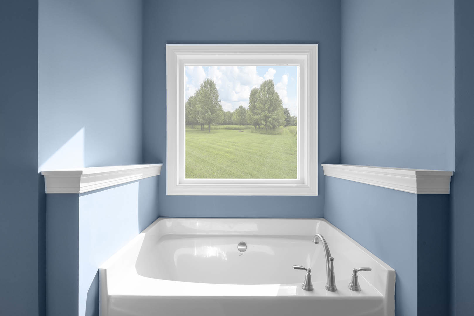 Freestanding white bathtub beneath large window overlooking green grass and trees, white wall molding, nearby sink with chrome faucet