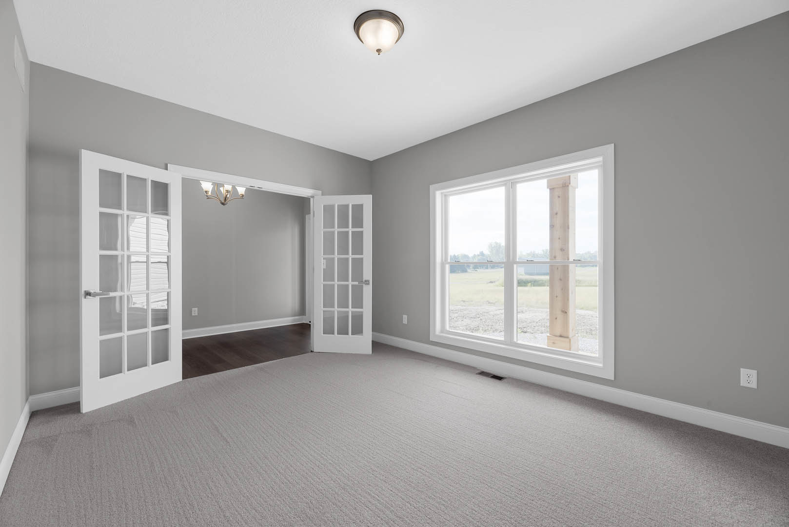 Spacious room with a large window framed by a white wooden pillar, glass-paneled white door, neutral carpet flooring, and ceiling light fixture.