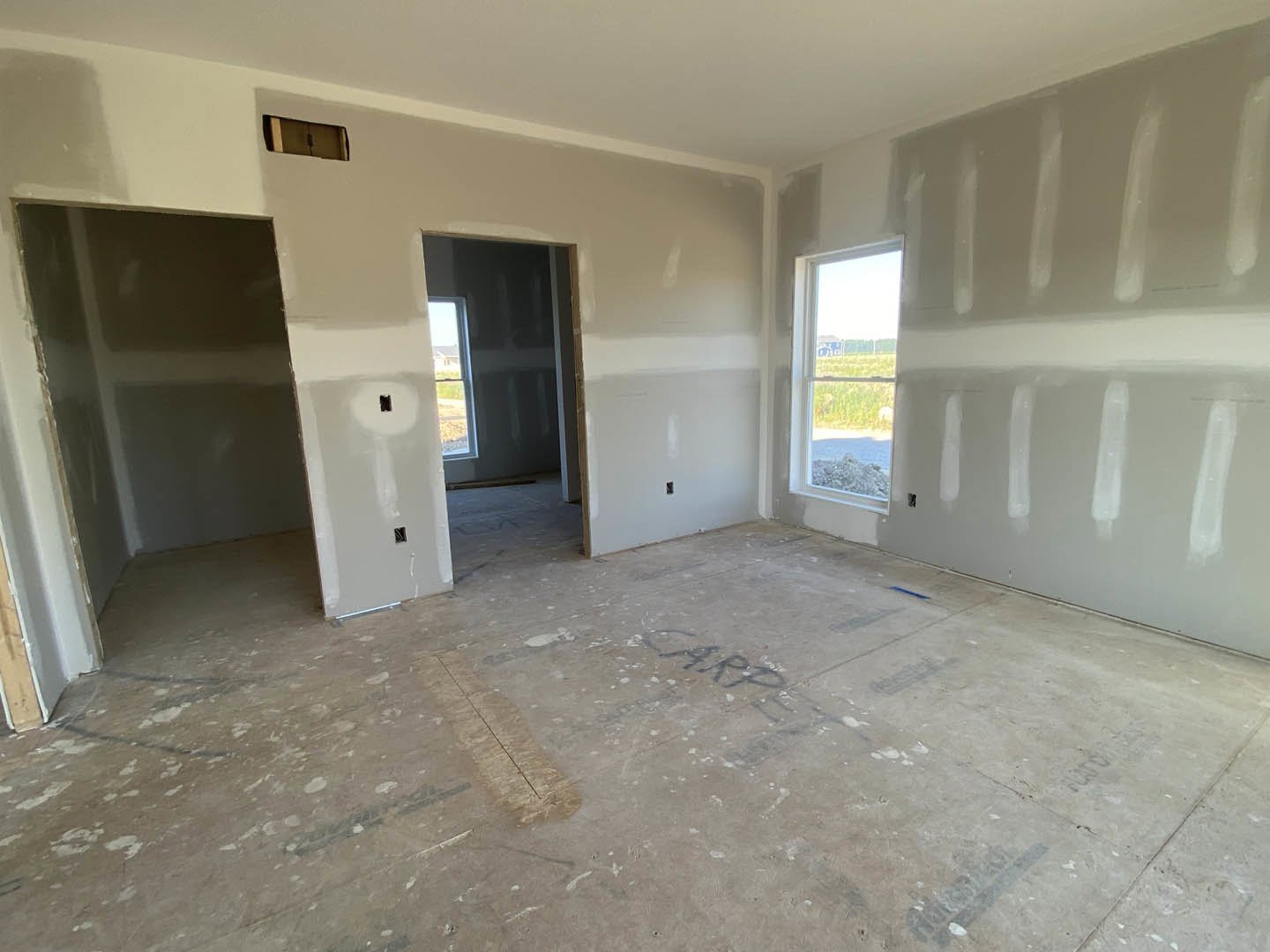 Bright room with smooth white plaster walls, large window overlooking grassy field, unfinished concrete floor with handwritten markings, simple white ceiling, and doorway leading
