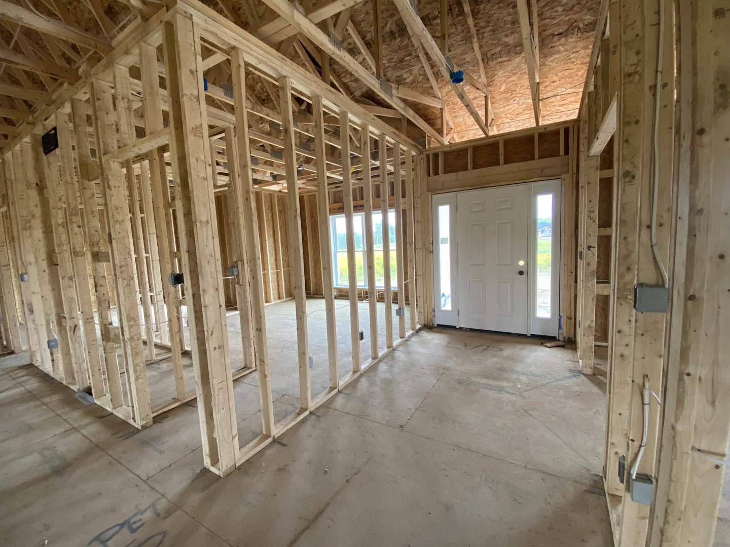 Wood-framed interior under construction with exposed beams, white door with glass panels, unfinished walls, and window overlooking green field