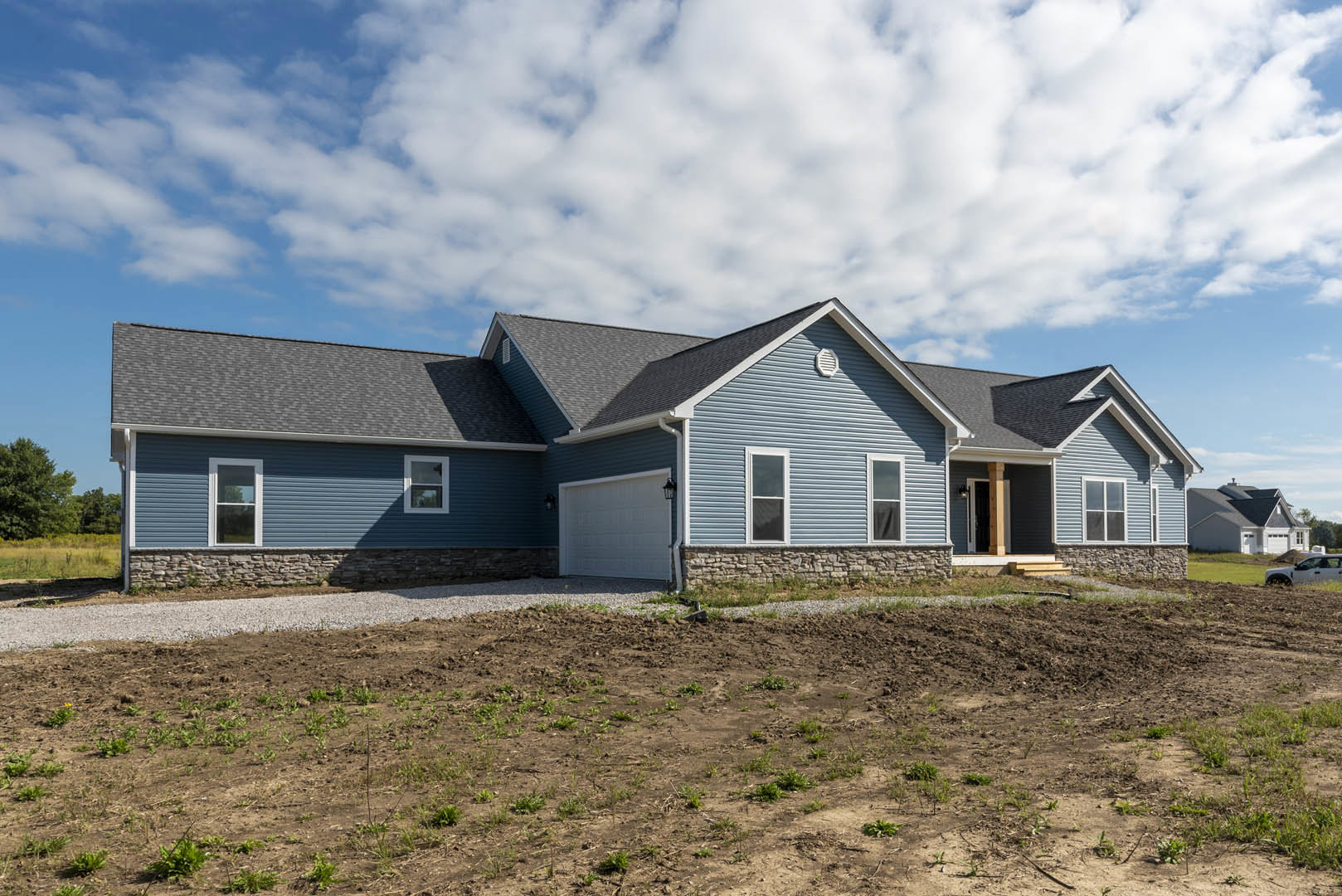 Two-story house with blue roof, white-framed windows, and attached garage, surrounded by a dirt field with scattered weeds under a cloudy sky