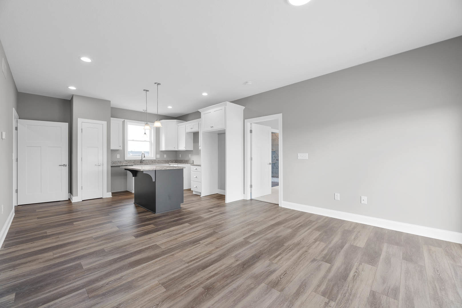 Open kitchen and dining area featuring hardwood floors, white walls, marble-topped kitchen island, white door with silver handle, large window, and modern light fixture.
