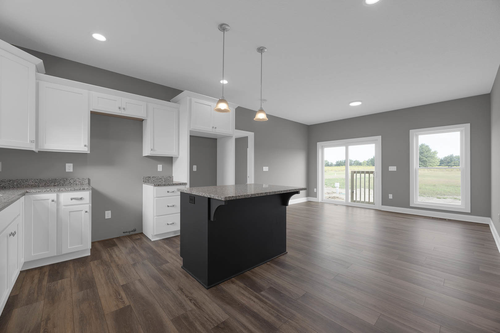 Kitchen with white cabinets and silver handles, black countertop island, wood flooring, window overlooking trees, sliding glass door with view of a grassy field, white door with