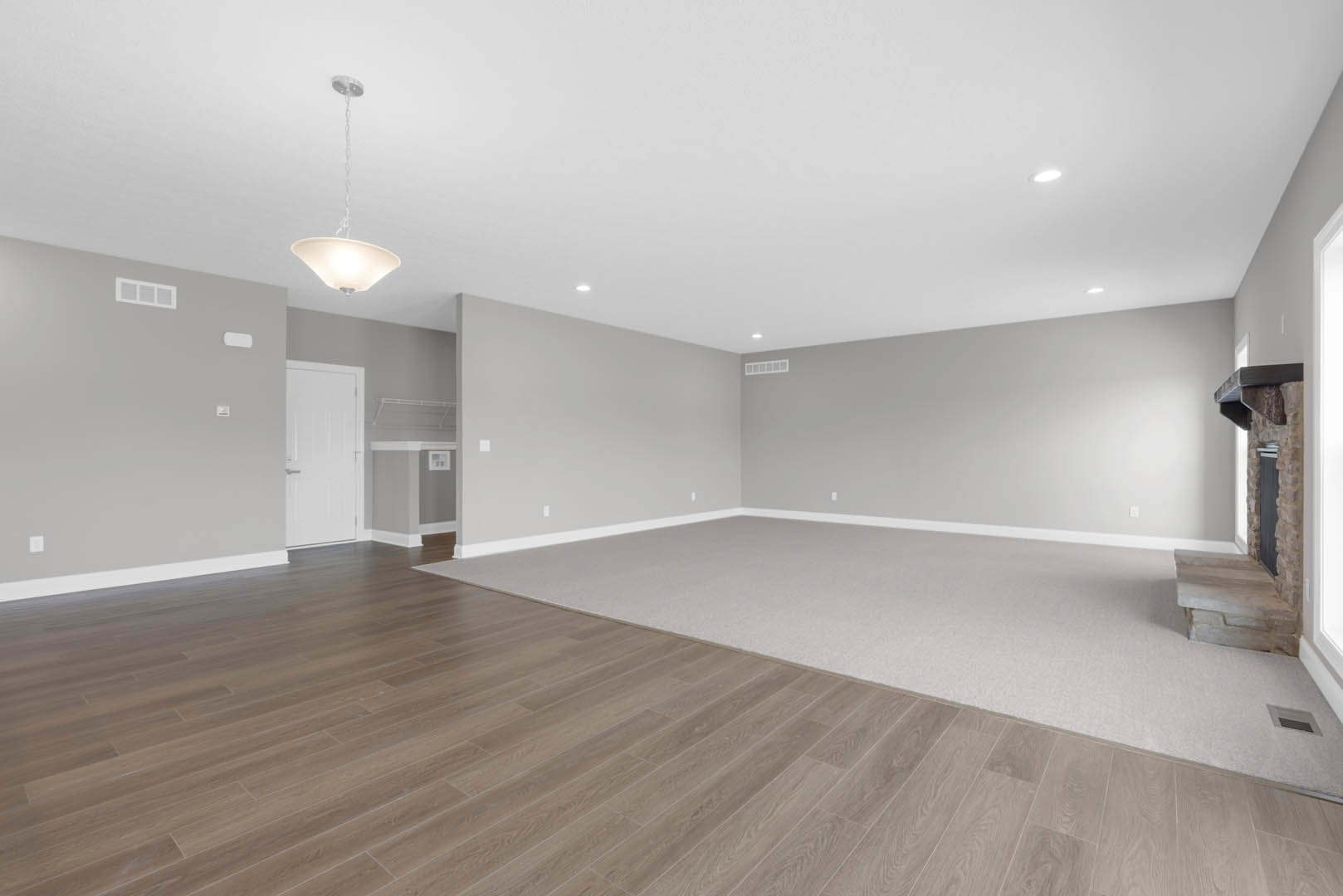 Spacious living area with hardwood flooring, white plaster ceiling, white door with metal handle, carpeted section, modern fireplace, recessed lighting, and decorative white bowl