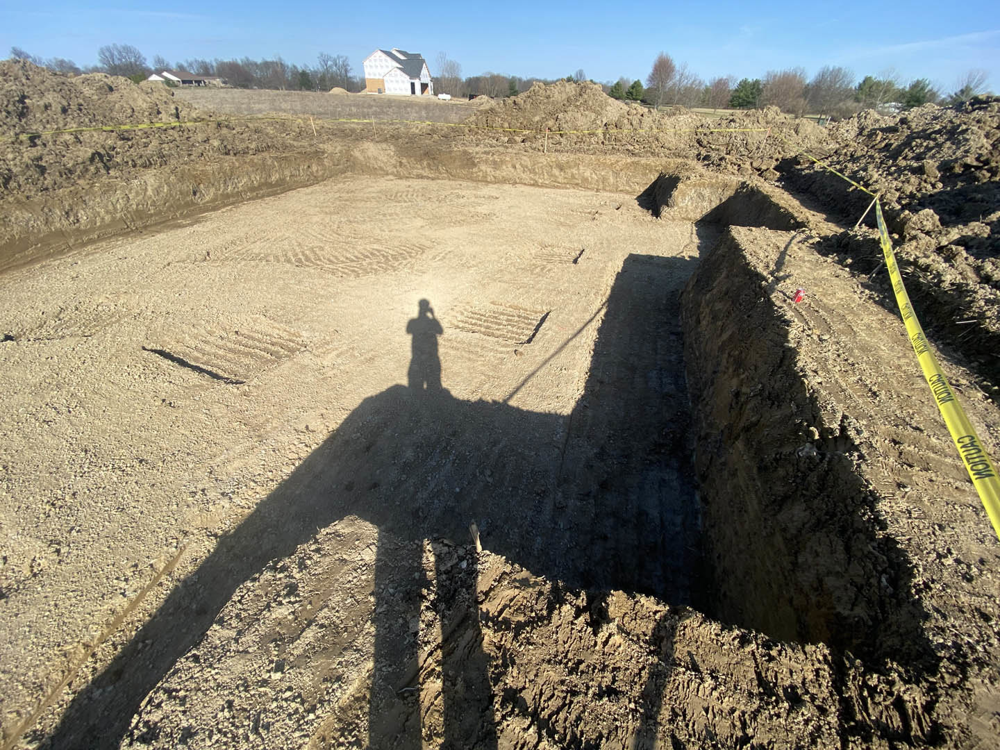 Shadow of a person cast on bare dirt in front of a custom home, yellow caution tape visible, blue sky and trees in background