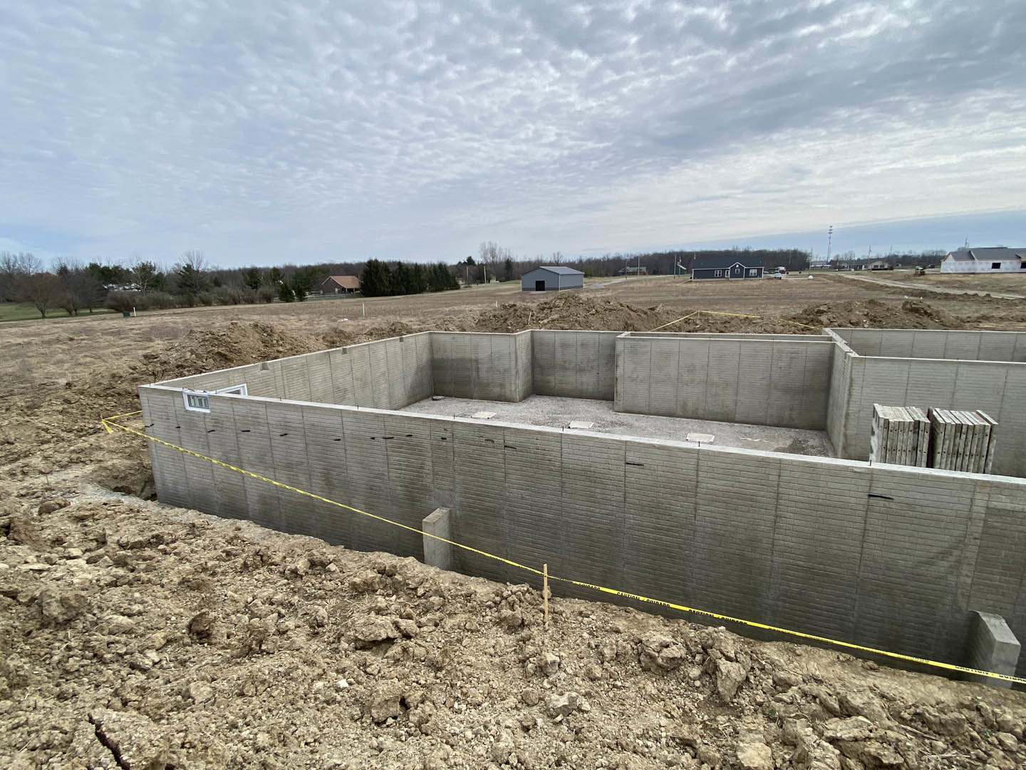 Concrete foundation and post surrounded by dirt and grass, yellow caution tape marking construction area, leafless tree and stacked wooden boxes nearby, cloudy sky overhead