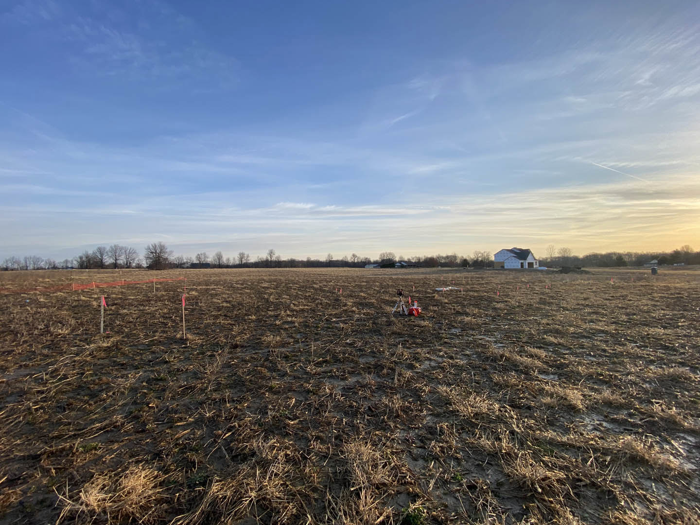 Dry grassy field with scattered orange poles, distant house partially obscured, blue sky with clouds overhead, lone tree near house, camera tripod visible in foreground