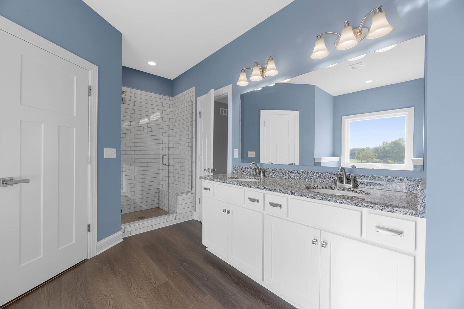 Bathroom with blue accent wall, white cabinets and drawers, white countertop, window showing trees outside, glass shower enclosure, white door with silver handle
