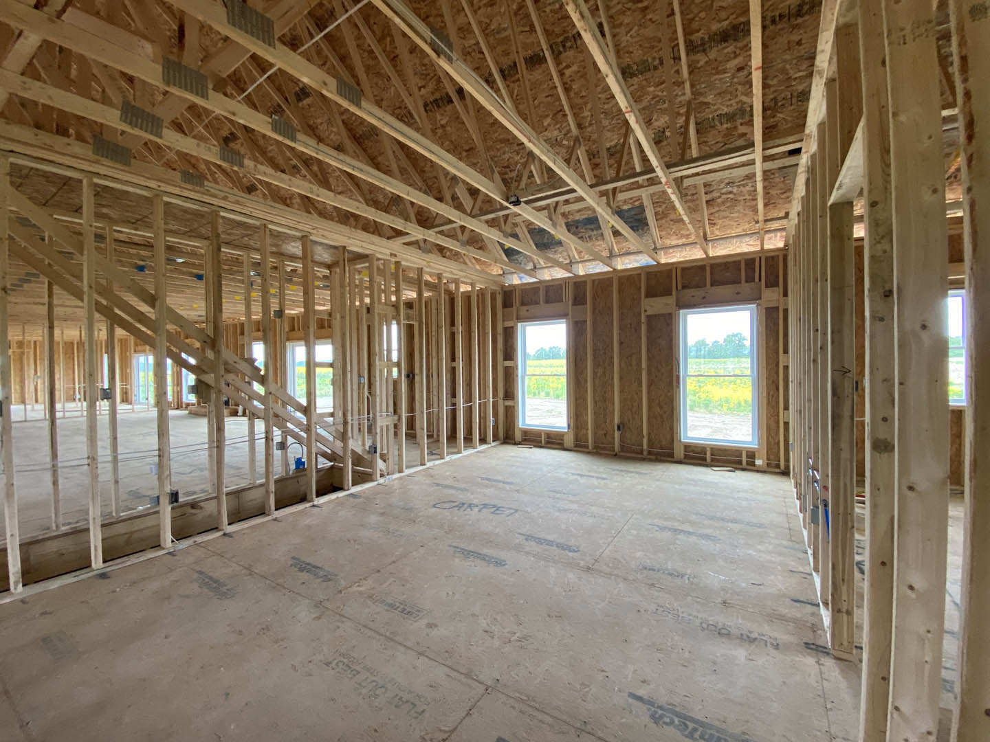Open living area with wood flooring, exposed ceiling beams, central staircase, and large windows overlooking a field of flowers