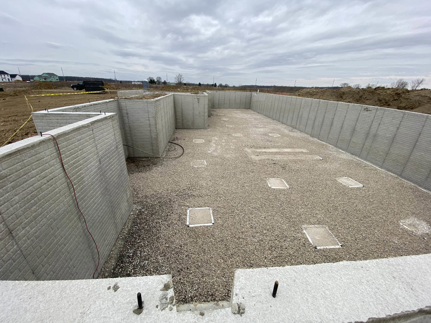 Concrete foundation with gravel fill, square blocks, red wire along wall, black truck parked in field, white square on dirt ground, cloudy sky overhead