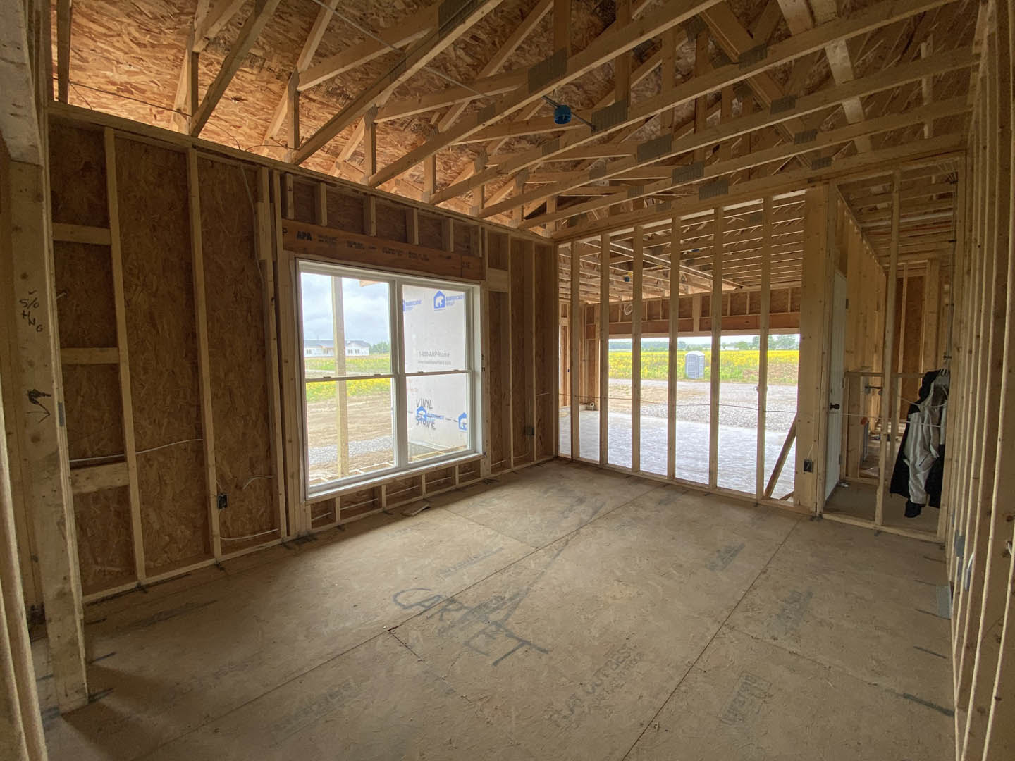 Sunlit room featuring a large window overlooking a grassy field, wood beam ceiling, light-colored flooring with visible markings, and neutral walls.